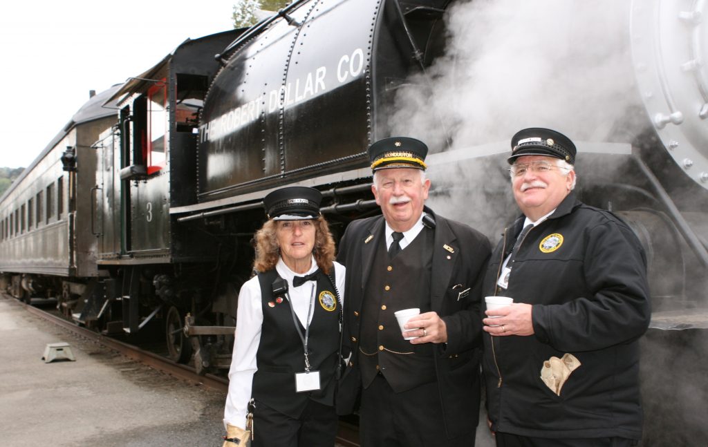 Two men and a woman in railroad conductors' uniforms who are part of the train crew pose in front of a steam locomotive.