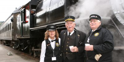 Two men and a woman in railroad conductors' uniforms who are part of the train crew pose in front of a steam locomotive.