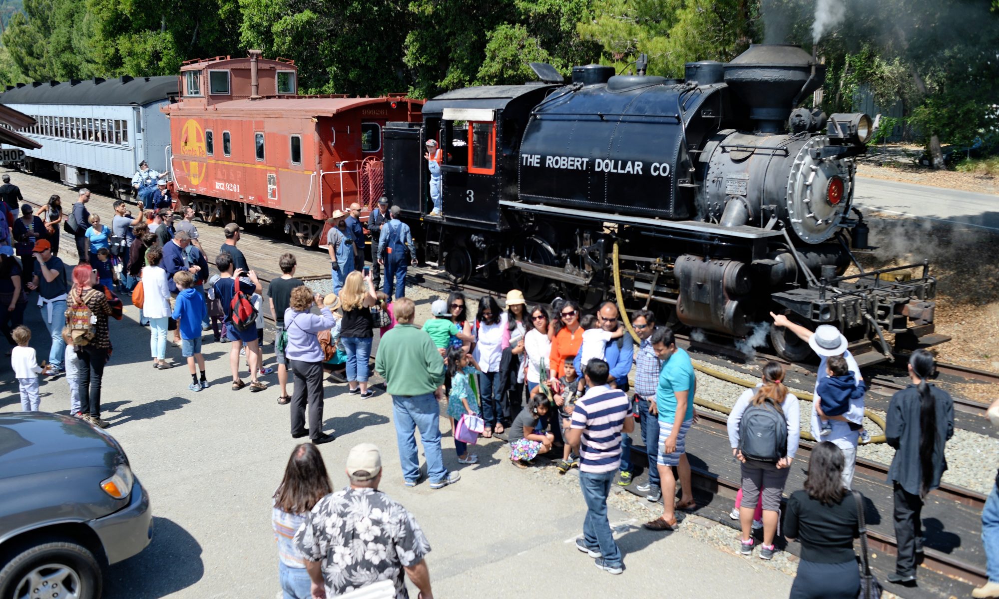 A steam locomotive powered passenger train sits in the Sunol station while a large group of passengers on the station platform view the locomotive and have their pictures taken in front of the locomotive.