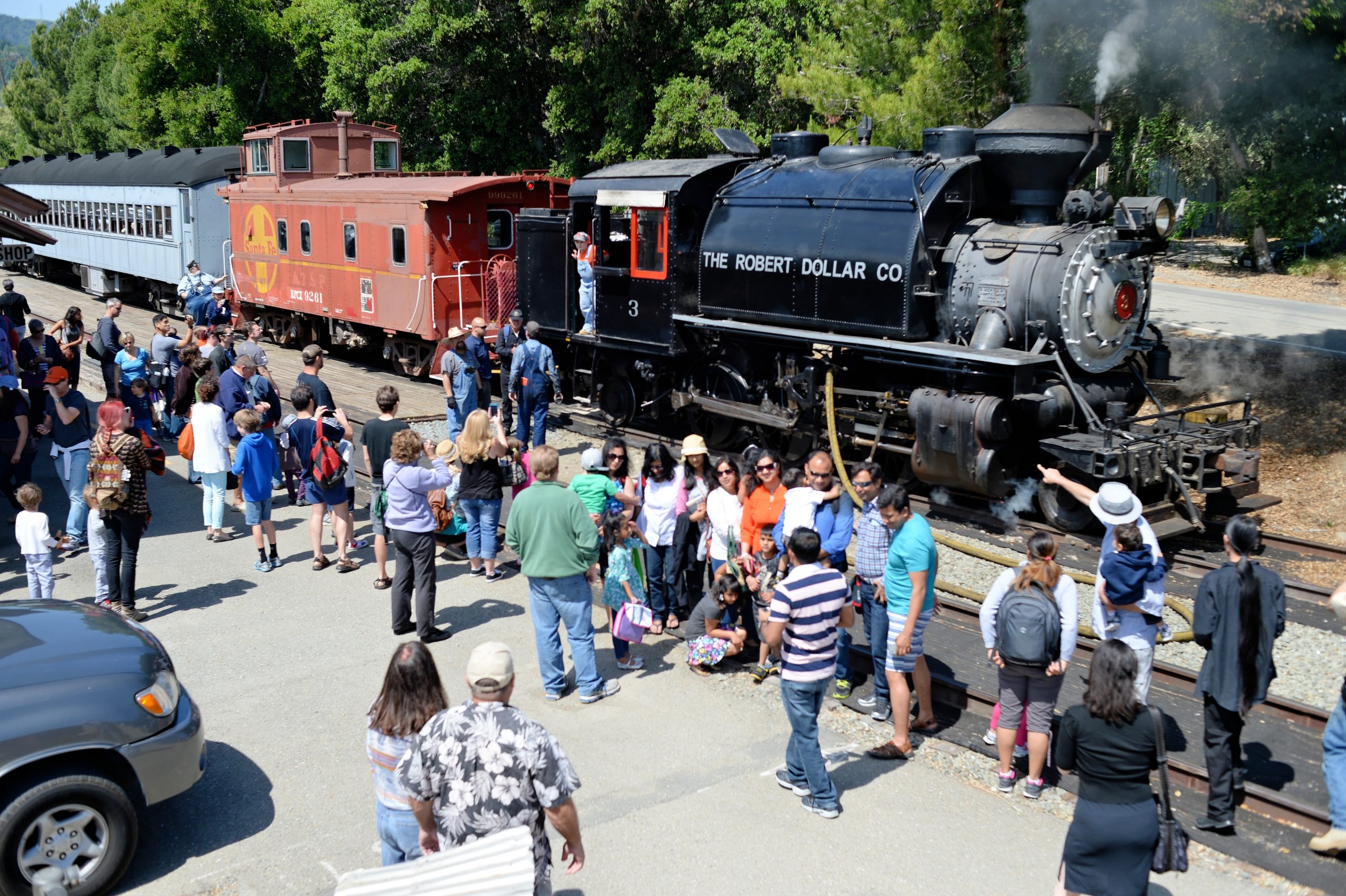 A steam locomotive powered passenger train sits in the Sunol station while a large group of passengers on the station platform view the locomotive and have their pictures taken in front of the locomotive.
