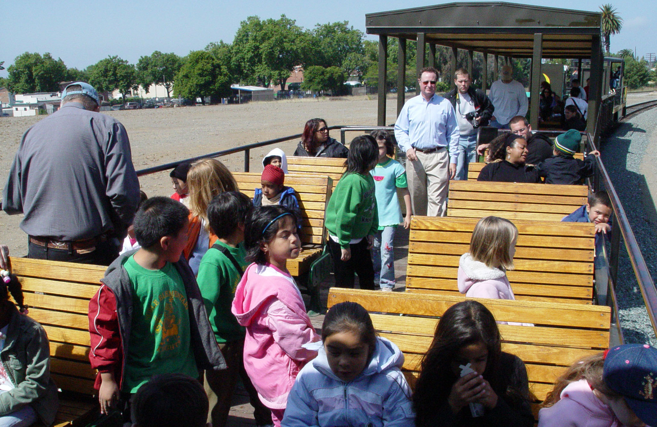 A photograph of a bunch of school kids in bench type seats that are part of an open air railroad passenger car as it waits for the train to depart from a railroad yard.
