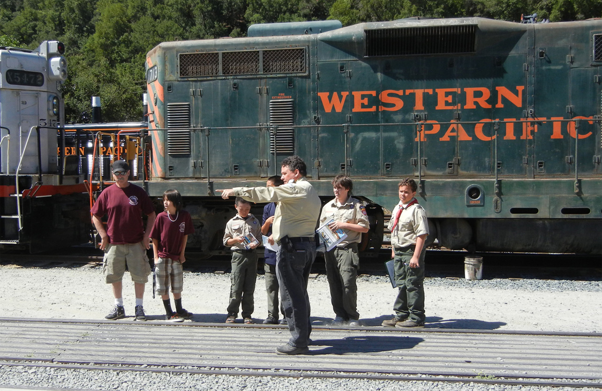 A Boy Scout Leader and museum volunteer points and explains different pieces of railroad equipment in the museum's railroad yard.