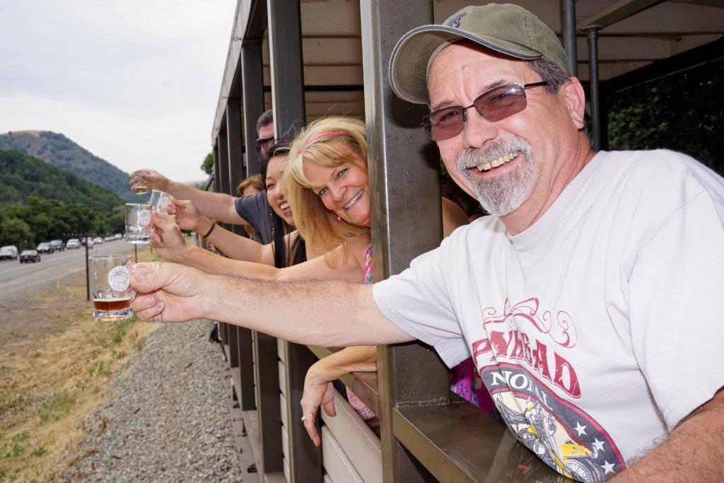 A photograph of several people riding on an open air railroad car where they are extending their arms out to display their small beer tasting glasses as the train runs along a highway.