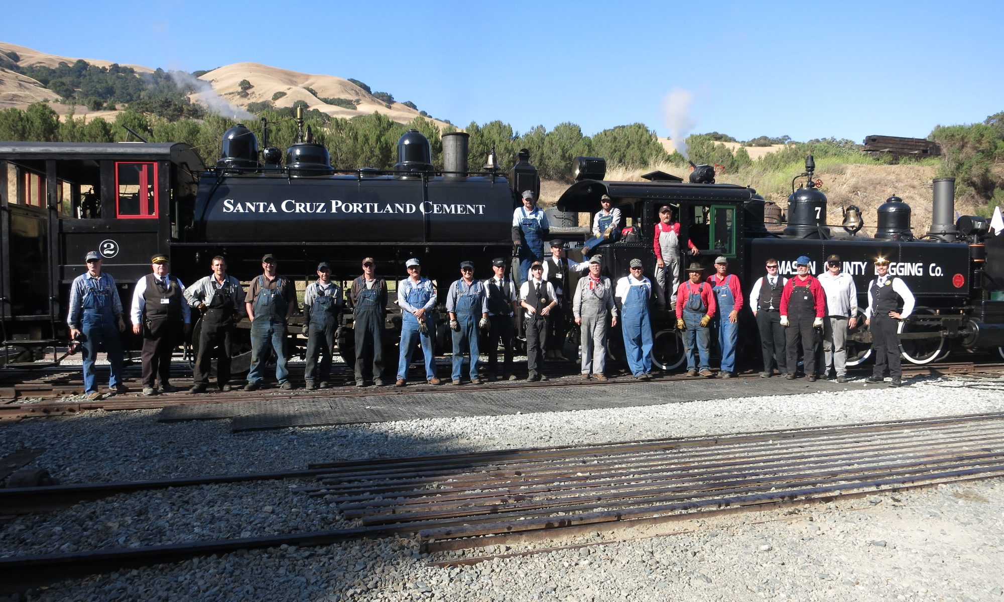 A large group of people most in railroad overalls standing in front of two steam locomotives