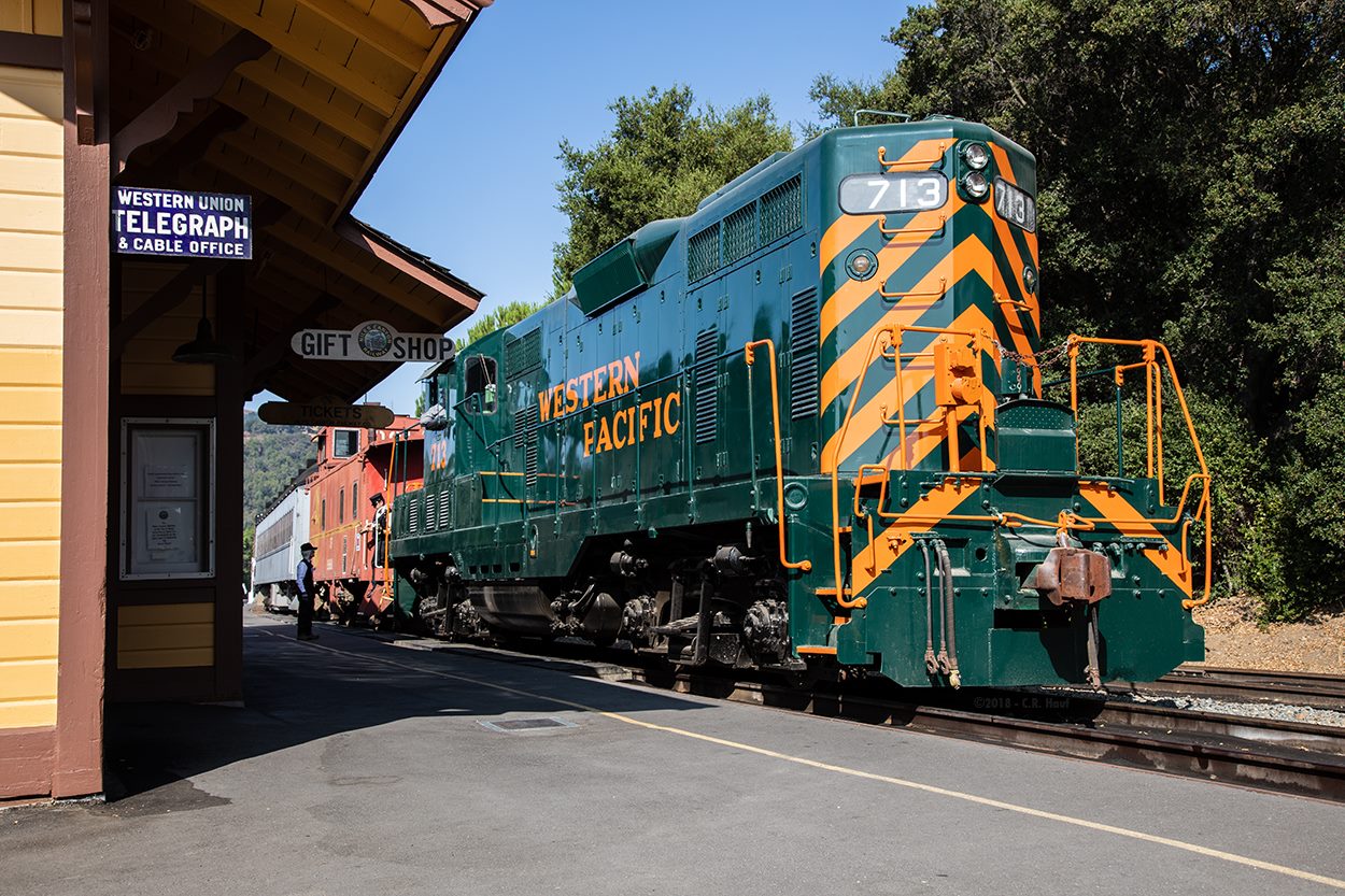 A historic freight diesel locomotive in the green and orange paint scheme of the Western Pacific Railroad sits in front of the museum's Sunol, California railroad station awaiting passengers.