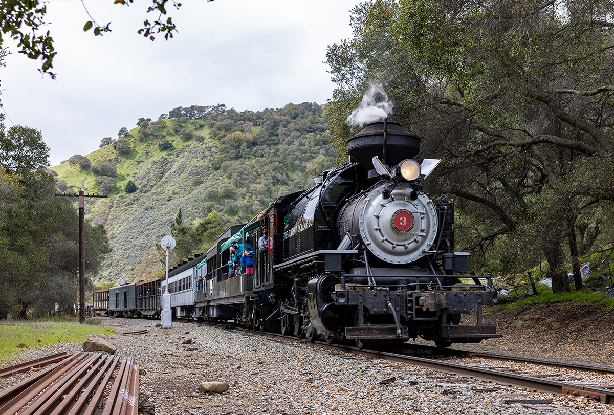 A steam locomotive pulling a passenger train rolls past a railroad signal on a part of the railroad deep inside of Niles Canyon.