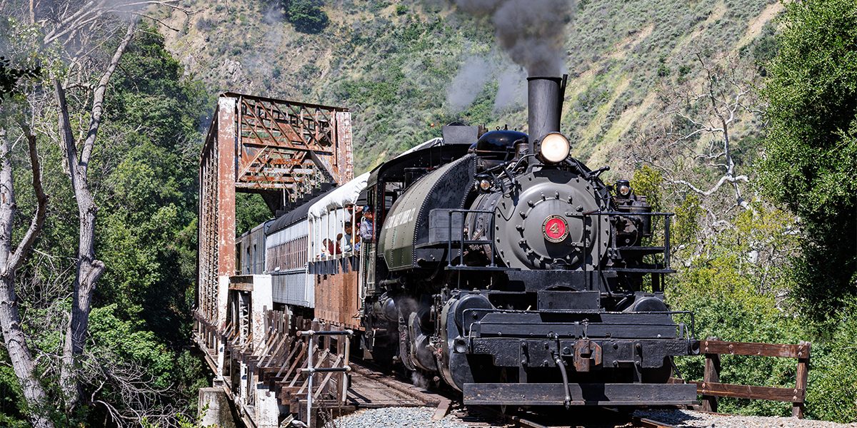 A steam locomotive pulling a passenger train over a steel bridge crossing a creek and located within a canyon.