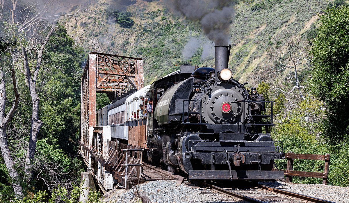 A steam locomotive pulling a passenger train over a steel bridge crossing a creek and located within a canyon.