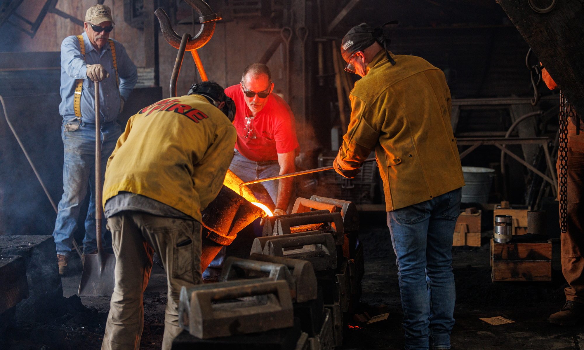 Three men in protective gear pour liquid iron from a ladle into a sand cast form sitting on the foundry floor with another man standing in the background with a shovel.