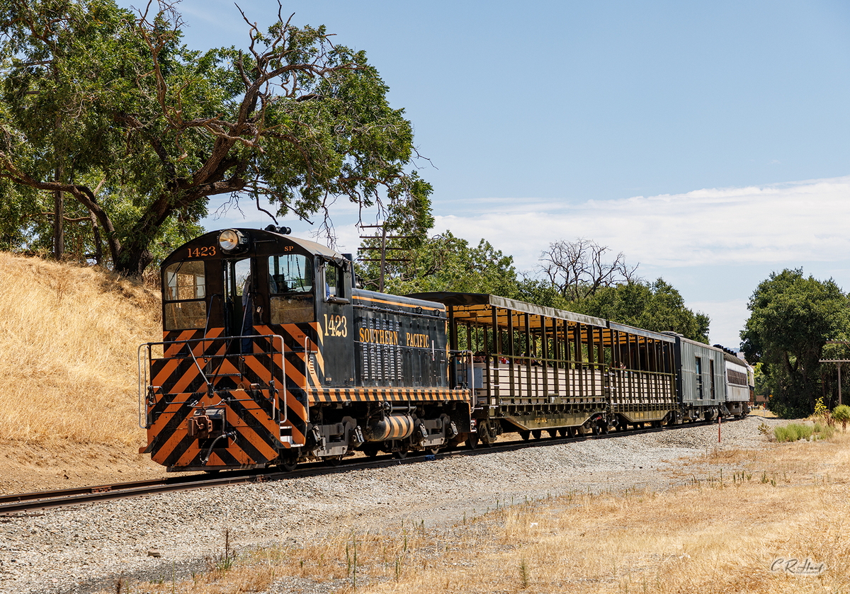 A historic diesel switcher locomotive pulls a passenger train along the tracks on a sunny California day.