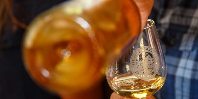 A close-up of a person's hand holding a wine glass with a blurry wine bottle pouring wine into the glass.