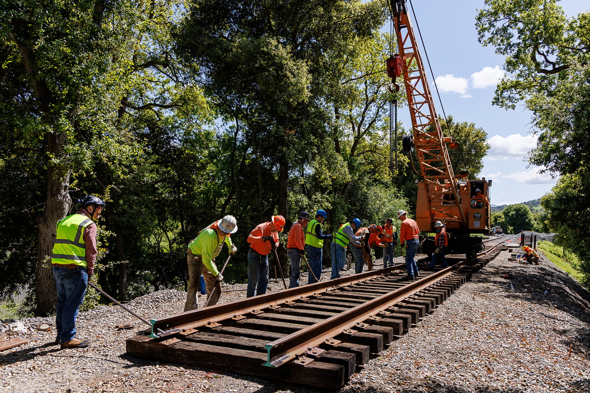A group of men and women in work clothes line up along a newly installed piece of railroad track as they work to properly align the track.