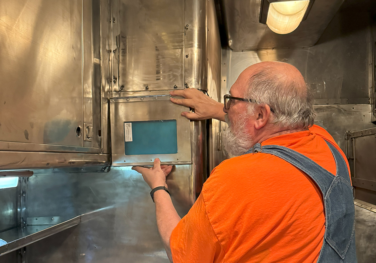 A man inside an all stainless steel railroad car kitchen holding up a stainless steel recipe book holder where it will be returned to its original mounted location.